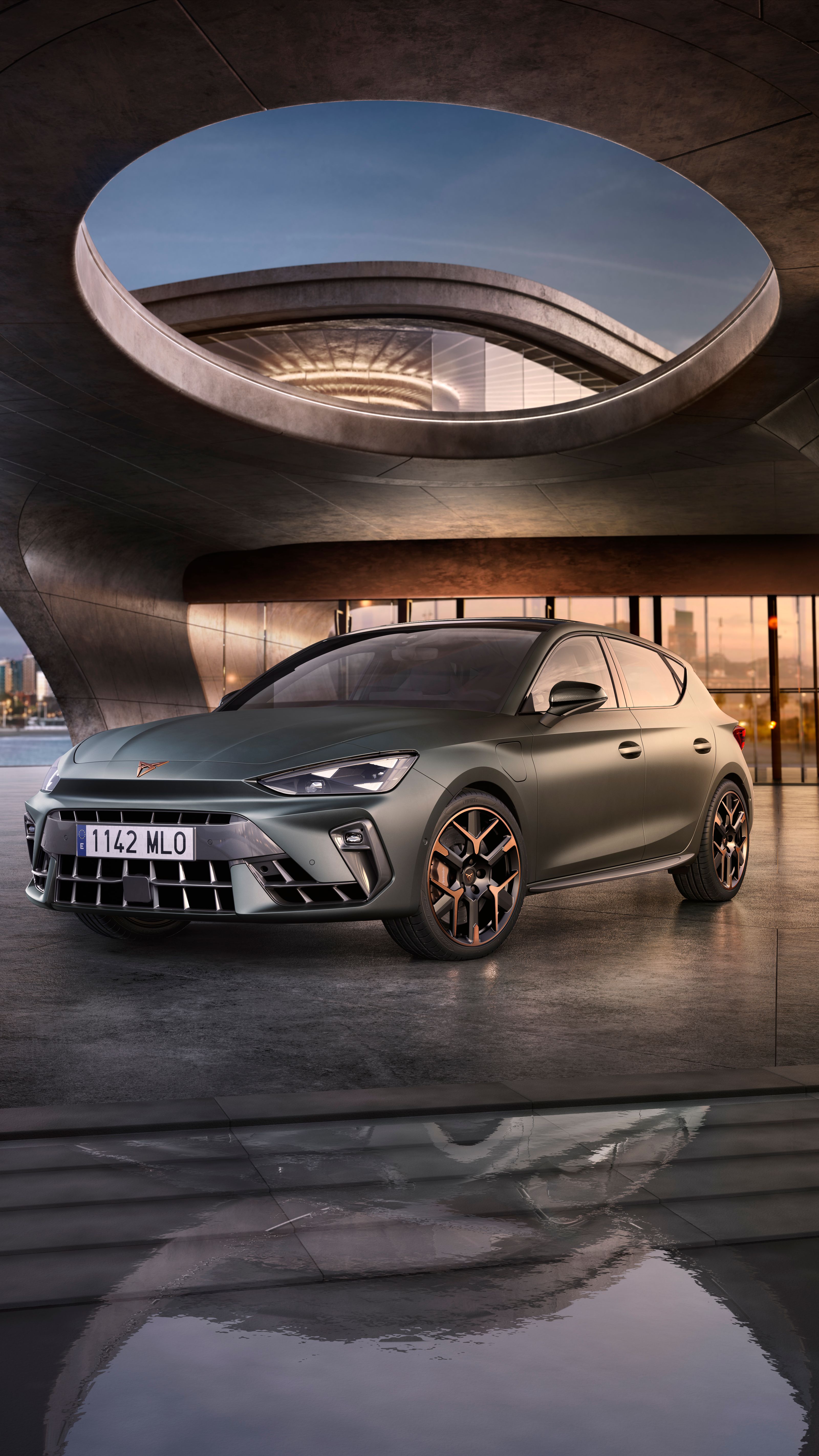 Automobil stojí na móle s modernou architektúrou v pozadí. Okolo je voda, na ktorej sa odráža okolie. V diaľke je silueta mesta.