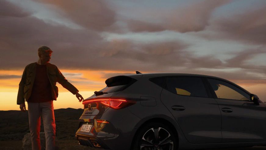 Muž stojí pri zaparkovanom sivom aute počas západu slnka. Pozadie tvorí dramatická obloha s farebnými mrakmi.