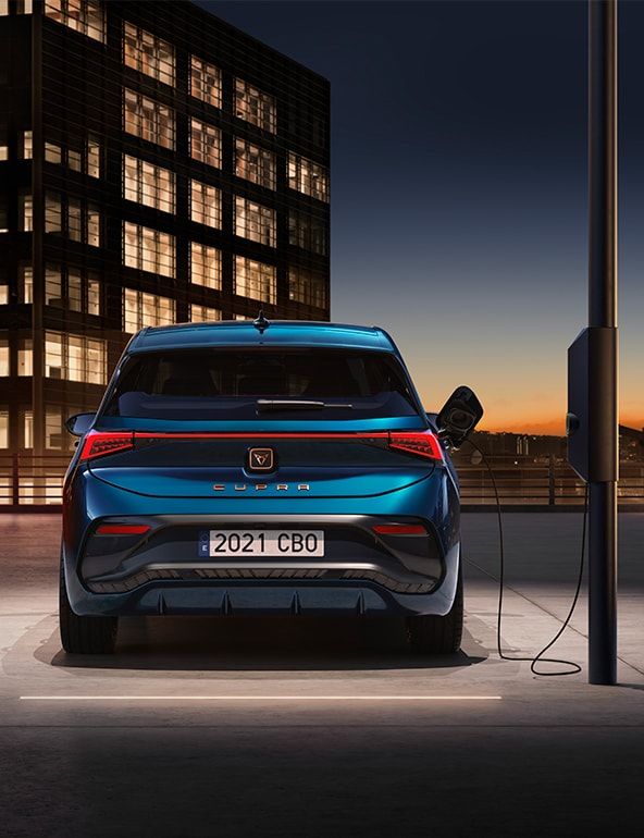Auto značky CUPRA je zaparkované pri nabíjacej stanici a je pripojené. Je to súčasť moderného mestského prostredia, ktoré zdôrazňuje technologický pokrok a udržateľnosť. Na pozadí je budova pri západe slnka.