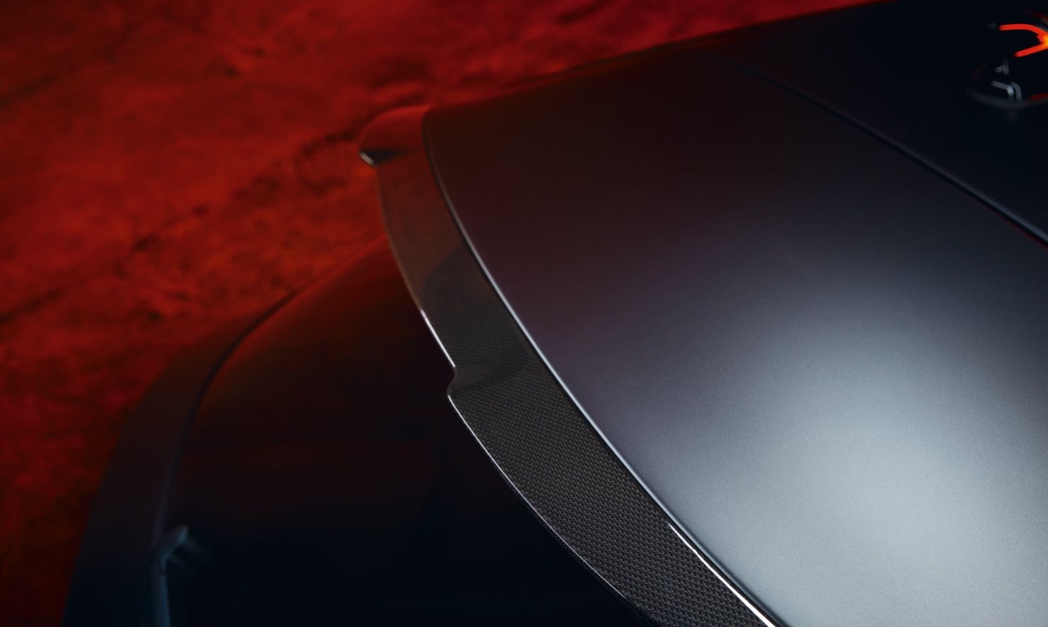 Detailný záber na strešný spojler športového auta, s dôrazom na jeho aerodynamický dizajn. Pozadie je osvetlené červeným svetlom, ktoré dodáva scéne dramatický efekt.