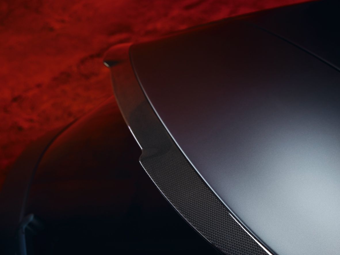 Detailný záber na strešný spojler športového auta, s dôrazom na jeho aerodynamický dizajn. Pozadie je osvetlené červeným svetlom, ktoré dodáva scéne dramatický efekt.