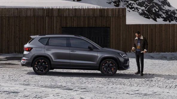 Šedé SUV CUPRA stojí pri drevenej budove v zasneženej horskej krajine. Muž stojí vedľa auta. V pozadí sú zasnežené hory.