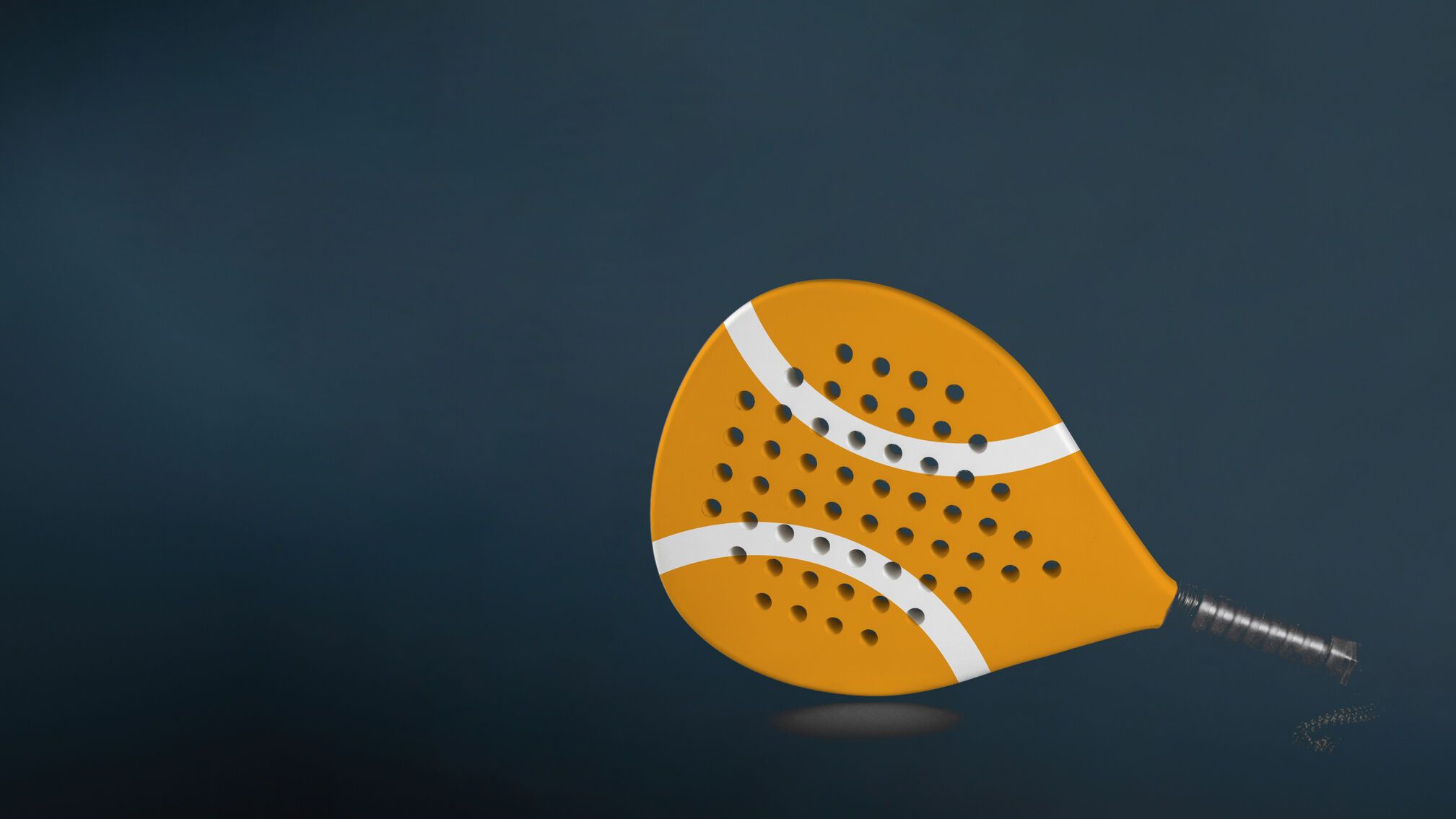 Na obrázku je oranžová padelová raketa s dvoma bielymi pruhmi. Raketa má veľa malých dierok po celej ploche. Je položená na tmavom pozadí.