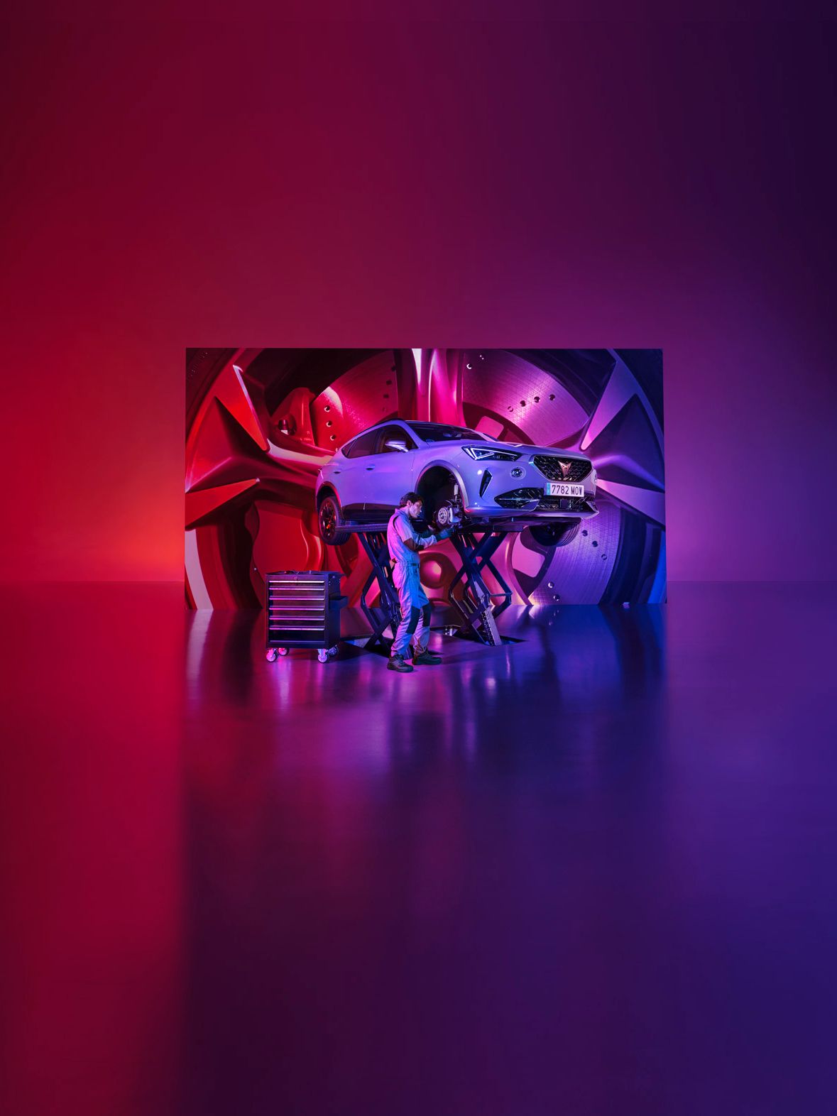 Auto zdvihnuté na zdviháku v servise, pozadie tvorí detail na koleso, dizajnový disk, brzdový kotúč a brzdový strmeň. Mechanik pri práci na aute, v popredí viditeľná skrinka s náradím. Celková atmosféra má moderný technologický vzhľad.