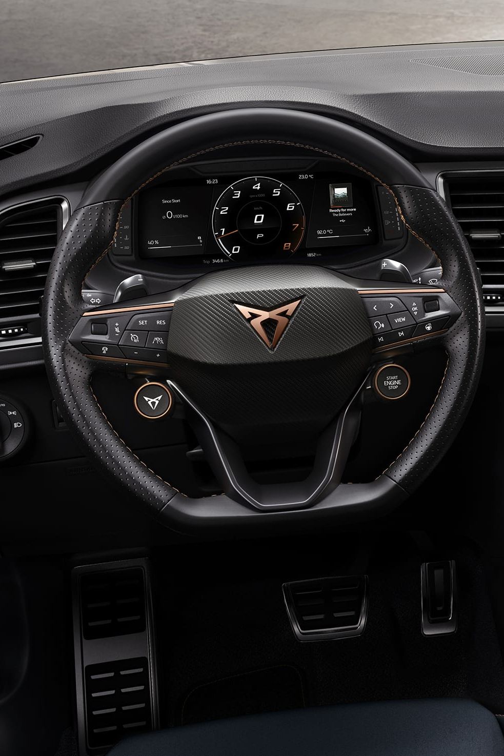 Multifunkčný volant automobilu s množstvom ovládacích prvkov a digitálny prístrojový panel za ním. V strede volantu je logo automobilovej značky. 