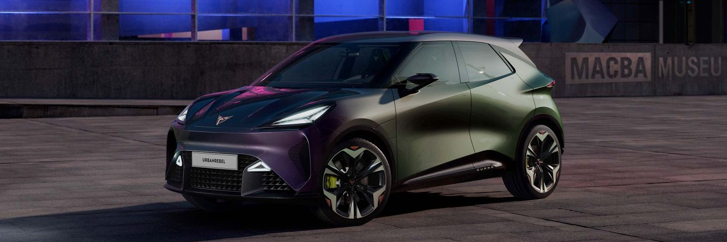 Moderné elektrické auto značky CUPRA zaparkovaný v nočnom meste. Auto má tmavú metalickú farbu a športový dizajn. V pozadí je mestská architektúra osvetlená farebnými svetlami.